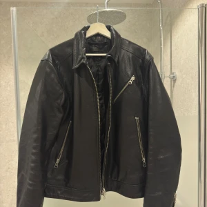 Svart skinnjacka Tiger of Sweden L - Riktigt snygg svart skinnjacka från Tiger of Sweden Jeans i storlek L. Jackan har flera silvriga dragkedjor, klassisk krage och quiltat innerfoder. Perfekt för dig som vill ha en stilren och edgy look.