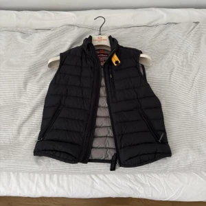 Svart dunväst från Parajumpers - Snygg svart dunväst från Parajumpers med quiltad design och dragkedja framtill. Västen har en cool gul detalj vid kragen och grått foder på insidan. Perfekt att slänga över en hoodie för extra värme. Flera praktiska fickor med dragkedja. Storlek 146-152