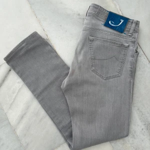Grå jeans från Jacob Cohen - Snygga grå jeans från Jacob Cohen med klassisk femficksdesign och ett unikt blått pälsmärke bak i midjan. Jeansen har raka ben och silverfärgade nitar, samt broderad J-logga på myntfickan. Insidan har coolt mönstrat tyg och inspirerande broderad text.