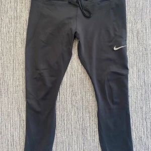 Svarta Nike tights med snörning - Svarta tights från Nike med smal passform och snörning i midjan. Swoosh-logga på vänster ben och paneler i stretchigt syntetmaterial för extra komfort. Perfekta för träning och har en sportig look.