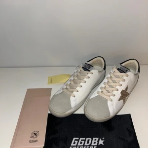 Golden Goose vita sneakers med glitter - Golden Goose sneakers i vitt skinn med beige snörning och grå mockadetalj på tån. Sidan har en glittrig guldstjärna och hälen är täckt av svart glitter med broderad logga. Platt sula och rund tå, riktigt snygga och unika detaljer. Helt nya och aldrig använda. Köpta som present men blev aldrig överlämnade, därför säljs de nu. 