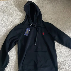 Svart hoodie från Polo Ralph Lauren - Svart hoodie från Polo Ralph Lauren med röd broderad logga på bröstet. Tröjan har huva med snörning och en stor magficka framtill. Mjuk och skön, perfekt för chill dagar. Klassisk stil med snygga detaljer och ribbade muddar.