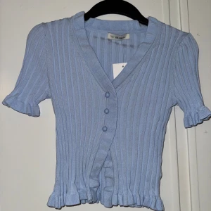 Ljusblå ribbad topp  - Superfin ljusblå ribbad topp från chiquelle  med v-ringning och tre knappar framtill. Kortärmad med volangdetaljer vid ärmslut och nederkant. Stickad känsla och slim passform, perfekt för dig som gillar söta detaljer och mjuka färger.