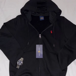 Svart Ralph Lauren full zip hoodie - Svart hoodie från Polo Ralph Lauren med dragkedja, huva och snörning. Klassisk röd broderad logga på bröstet. Tillverkad i mjuk bomull, perfekt för chill dagar. Snygg och enkel att matcha med jeans eller joggers.