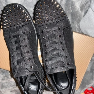 Svarta Christian Louboutin sneakers med nitar - Säljer ett par svarta sneakers från Christian Louboutin med glittrig finish och coola nitar över hela skon. Skorna har snörning, rund tå och den klassiska röda sulan med Louboutin-logga. Materialet är skinn och syntet, och de har platt sula.