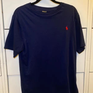 Mörkblå t-shirt från Polo Ralph Lauren - Snygg mörkblå t-shirt från Polo Ralph Lauren med röd logga på bröstet. T-shirten har rund hals och korta ärmar, tillverkad i mjuk bomull som känns skön mot huden. Perfekt för dig som gillar stilrena och grish plagg. Den är väldigt stor i storleken så därför så passar den S och i vissa fall till och med M.