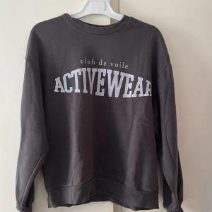 Mörkgrå sweatshirt från Gina Tricot - Mörkgrå sweatshirt från Gina Tricot med trycket 'club de voile ACTIVEWEAR' i vitt framtill. Tröjan har rund hals, ribbade muddar vid ärmslut och nederkant samt en loose passform. I storlek S men kan passa dig som har större
