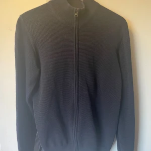 Mörkblå stickad kofta från Signal - Mörkblå stickad kofta från Signal med hel dragkedja framtill och hög krage. Tröjan har en clean look med ribbade muddar och lång ärm, perfekt för lager-på-lager. Klassisk och enkel design som funkar till det mesta. Använd fåtal gånger