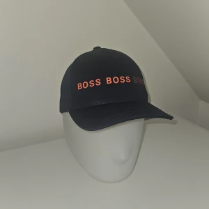 Svart keps från BOSS med orange text - Snygg svart keps från BOSS med justerbar rem bak och orange text 'BOSS' tryckt flera gånger framtill. Klassisk böjd skärm och ventilerande hål upptill. Perfekt accessoar för en cool och sportig look. Aldrig använd nästan