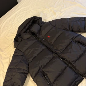 Svart pufferjacka från Polo Ralph Lauren - Svart pufferjacka från Polo Ralph Lauren med röd broderad logga på bröstet. Jackan har huva, två dragkedjeförsedda fickor och quiltad design. Perfekt för kalla dagar och har en clean, sportig look. Använd någon enstaka gång. Pris kan diskuteras😁