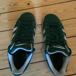 Gröna sneakers med vita detaljer - Snygga gröna sneakers med vita ränder på sidorna och vit logga på plösen. Skorna har klassisk snörning och en rund tå. Insidan är fodrad i vitt och de har en sportig vibe som passar perfekt till streetwear.