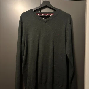 Mörkgrå ulltröja Tommy Hilfiger L - Snygg mörkgrå stickad tröja från Tommy Hilfiger i tvättbar ull. Klassisk v-ringning och diskret logga broderad på bröstet. Perfekt för dig som gillar stilrena och tidlösa plagg med premiumkänsla.