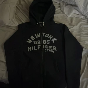 Svart hoodie från Hilfiger Denim M - Svart hoodie från Hilfiger Denim med tryck 'New York US 85 Hilfiger Denim' på bröstet. Tröjan har dragkedja framtill, huva och långa ärmar. Perfekt för en chill och avslappnad stil.