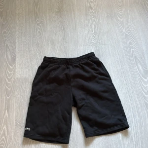 Svarta shorts från Lacoste Sport - Svarta shorts från Lacoste Sport med klassisk krokodillogga broderad på benet. De har elastisk midja och är gjorda i mjuk bomull, perfekta för chill eller sport. Passformen är normal och längden går till knäna. Snygg och enkel design som funkar till allt.