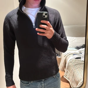 Svart stickad half zip-tröja M från A Day’s March - Svart stickad tröja från A days MARCH i storlek M. Tröjan har en hög, ribbad krage och en snygg half zip-dragkedja framtill. Perfekt till jeans och funkar året runt när du vill ha en clean och stilren look.