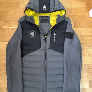 Sailracing hybridjacka - grå och svart dunjacka från Sail Racing med gul insida .                                                                         Fylld med högkvalitativt dun från Allied Feather & Down. Perfekt för kalla dagar och snygg streetstil. 