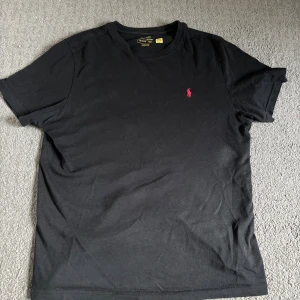 Svart t-shirt från Polo Ralph Lauren - Svart t-shirt från Polo Ralph Lauren med klassisk röd broderad logga på bröstet. T-shirten är i bomull och har en normal passform med korta ärmar och rund hals. Perfekt basic-plagg med stilren look.