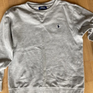 Grå sweatshirt från Polo Ralph Lauren - Klassisk grå sweatshirt från Polo Ralph Lauren med rund halsringning och broderad marinblå logga på bröstet. Tröjan har långa ärmar och ribbade muddar vid ärmslut och nederkant. Perfekt för en avslappnad och stilren look. Storlek passar lilla 13-17 år eller XS herr. Passar längd 160-168