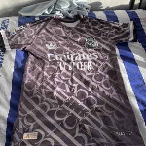 Real Madrid Mbappé fotbollströja - Säljer en snygg Real Madrid fotbollströja från Adidas med Mbappé och nummer 9 på ryggen. Tröjan är mörkgrå med ett unikt mönster och vita detaljer, samt tryck från Emirates och klubbmärket på bröstet. Tillverkad i lätt och ventilerande material.