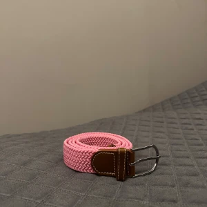 Flätat rosa bälte med skinn-detaljer - Snyggt flätat bälte i rosa med bruna skinn-detaljer och silvrigt spänne. Bältet har en flexibel och mjuk känsla tack vare det flätade materialet och passar perfekt för att ge outfiten lite extra färg. Stilren design med kontrastsömmar på skinnet.