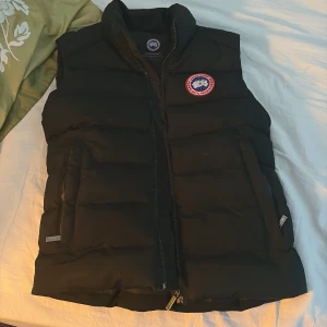 Svart dunväst från Canada Goose - Svart dunväst från Canada Goose med klassisk logga på bröstet. Västen har hög krage, två fickor med dragkedja och quiltad design.OBS ett hål i höger ficka så lägg inget litet där som lätt kan föesvinna!  Perfekt för lager-på-lager och håller dig varm under kyliga dagar.