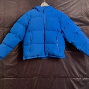 Blå pufferjacka från Lacoste - Snygg blå pufferjacka från Lacoste med huva och dragkedja framtill. Jackan har två fickor med dragkedja och en stor broderad krokodil på ryggen. Perfekt för kalla dagar när du vill hålla stilen och värmen.