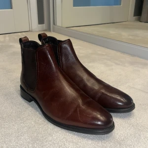 Bruna Chelsea boots från Hush Puppies - Snygga Chelsea boots i mörkbrunt skinn från Hush Puppies. Klassisk rund tå, elastiska paneler på sidorna och låg klack. Perfekt för dig som gillar stilrena och tidlösa boots med en clean look. Säljes i originalkartong. Skorna är mycket endast testade aldrig använda utomhus. Nypriset ligger på 1099kr