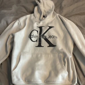 Vit hoodie från Calvin Klein Jeans - Vit hoodie från Calvin Klein Jeans med stor svart CK-logga framtill. Klassisk känguruficka och huva. Perfekt för dig som gillar stilrena och trendiga plagg med tydlig logga. Mjuk och skön, passar till allt.