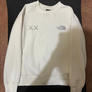 North face x kaws tröja - Säljer en clean vit sweatshirt från The North Face med broderad logga och två X-detaljer på bröstet. Tröjan har rund hals, långa ärmar och ribbade muddar. Perfekt för dig som gillar stilren och enkel streetwear.