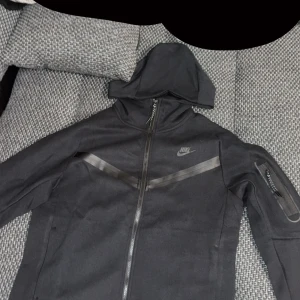 Svart vindjacka från Nike med huva - Svart Nike vindjacka med huva och hel dragkedja framtill. Snygga svarta detaljer i form av diagonala ränder över bröstet och en ficka med dragkedja på ärmen. Jackan har en sportig look och är perfekt för dig som gillar streetwear. PRIS KAN DISKUTERAS!