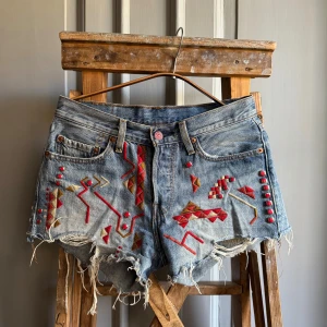 Levi's denim shorts med broderi st 26  - Unika tribal Levi's 501 jeansshorts med broderi samt röda nitar. Klassisk femficksmodell i ljusblå denim med slitna detaljer och fransig nederkant. St 26 