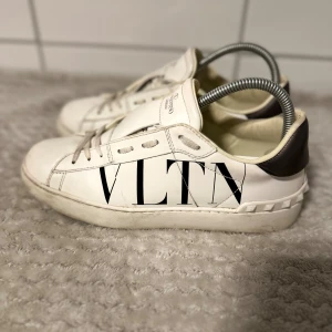 Valentino VLTN vita sneakers i skinn - Snygga vita sneakers från Valentino med VLTN-logga på sidan. Skorna har svarta detaljer på hälen och coola nitar runt sulan. Tillverkade i skinn med klassisk snörning och rund tå. Perfekta för dig som vill sticka ut med en lyxig streetstyle.