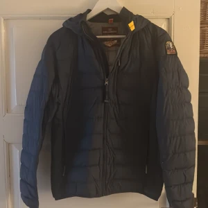 Mörkblå lättviktsjacka Parajumpers - Snygg mörkblå lättviktsjacka från Parajumpers med huva och klassisk PJS-patch på ärmen. Jackan har quiltad design, dragkedja framtill och cool gul detalj vid kragen. Perfekt för dig som vill ha en stilren och funktionell jacka.