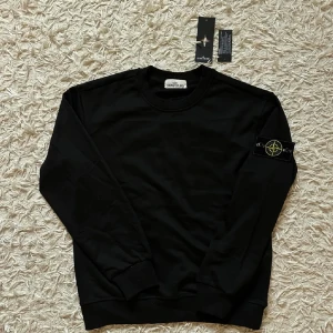 Svart sweatshirt från Stone Island - Svart sweatshirt från Stone Island med klassisk rund hals och ribbade muddar. Tröjan har den ikoniska loggan på vänster ärm i gult och grönt. Tillverkad i mjuk bomull för en skön och stilren look.