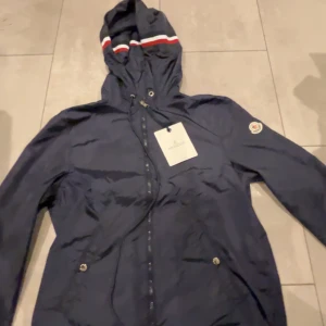 Marinblå vindjacka från Moncler - Snygg marinblå vindjacka från Moncler med huva och dragsko. Jackan har dragkedja framtill, två fickor med tryckknappar och Moncler-logga på ärmen. Huvan har coola ränder i vitt och rött. Perfekt för dig som vill ha en stilren och sportig look. Du får till snöret . Size M