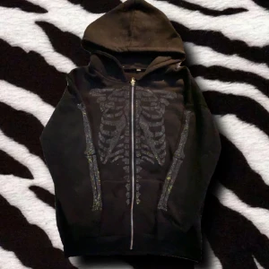 Svart hoodie med skelettmönster - Cool svart hoodie med glittrigt skelettmönster på framsidan. Tröjan har dragkedja hela vägen, huva och långa ärmar. Perfekt för dig som gillar edgy stil och vill sticka ut lite extra.