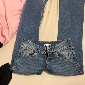 Blå jeans med broderade rosetter - Snygga blå jeans från Gina Tricot med låg midja och bootcut. Jeansen har broderade rosetter och detaljer på bakfickorna, samt råa, fransiga benslut och dubbla knappar fram. Perfekt för dig som gillar unika detaljer och en avslappnad stil.