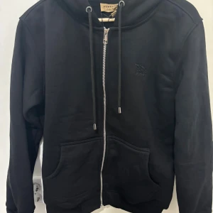 Svart Burberry kofta - Svart Burberry med hel dragkedja, snörning i huvan och klassiskt rutigt foder på insidan. Tröjan har två stora fickor framtill och diskret broderad logga på bröstet. Perfekt för dig som gillar stilrena och exklusiva plagg.