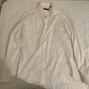 Vit skjorta från Dressmann i bomull - Klassisk vit skjorta från Dressmann i premium bomull. Skjortan har lång ärm, knappar framtill och en stilren krage. Perfekt för dig som gillar en clean och enkel look.