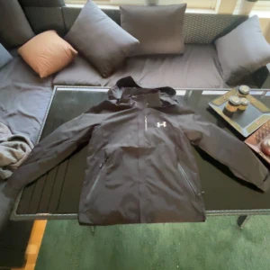 Svart vindjacka från Under Armour - Svart vindjacka från Under Armour med huva och dragkedja framtill. Jackan har två sidofickor med dragkedja och ett stilrent vitt logotryck på bröstet. Materialet är lätt och syntetiskt, perfekt för blåsiga dagar. Snygg och sportig look med normal passform.