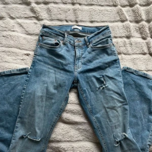 Ljusblå slitna bootcut jeans - Säljer ett par ljusblå jeans med bootcut-modell och slitningar på båda benen. Jeansen har klassisk femficksdesign och är tillverkade i denim. Perfekta för dig som gillar en avslappnad och trendig look.