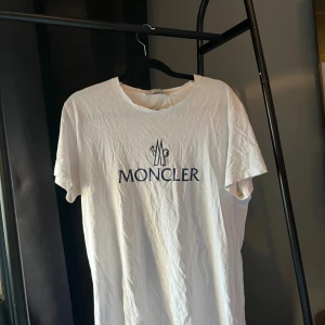 Vit Moncler t-shirt med logga - Vit t-shirt från Moncler i bomull med klassisk rund hals och stor mörkblå Moncler-logga framtill. Enkel och stilren design, perfekt för dig som gillar exklusiva märken och clean look.