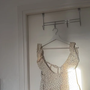 Prickig sommarklänning med volang - Supersöt ljus topp med svarta prickar och volangdetaljer vid axlarna. Som har en öppen rygg med knytband och är ärmlös, perfekt för varma dagar. Den har en luftig passform och ger en romantisk vibe.