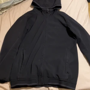 Svart vindjacka från Sail Racing - Svart vindjacka från Sail Racing med huva och diskret logga på bröstet. Jackan har dragkedja framtill, två sidofickor och ribbade muddar med gråa detaljer. Materialet är slitstark polyester och passformen är normal. Perfekt för dig som gillar stilren och sportig look.