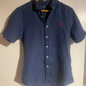 Blå linneskjorta från Ralph Lauren - Snygg blå kortärmad skjorta från Ralph Lauren i slim fit. Tillverkad i linne med klassisk knappslå och röd broderad logga på bröstet. Perfekt för en stilren och fräsch look under varmare dagar.