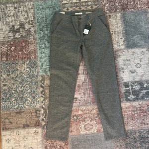 Grå chinos från Hansen & Jacob - Snygga grå chinos från Hansen & Jacob i klassisk modell med raka ben och diskreta fickor. Byxorna har bälteshällor, knapp och dragkedja fram samt bakfickor med knapp. Materialet är mjukt och ser ut att vara bomullsmix, perfekt för en clean och stilren look.