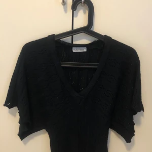 Svart stickad topp från Clockhouse - Svart stickad topp från Clockhouse med v-ringning och korta ärmar. Toppen har ett diskret hålmönster och är figurnära nertill. Perfekt för dig som gillar en enkel men ändå snygg stil.