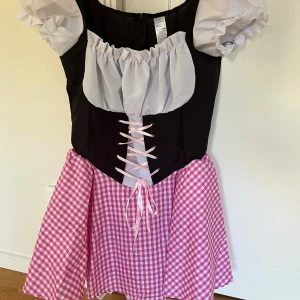 Oktoberfest klänning - Supergullig kortklänning i rosa och vitt med rutigt mönster, puffärmar och snörning framtill. Klänningen har svart liv, vit volang upptill och dragkedja bak. Perfekt för oktoberfesten!