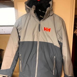  Skidset från Helly Hansen - Säljer en snygg tvåfärgad skidset från Helly Hansen. Jackan har huva, dragkedja framtill och två fickor med dragkedja. Röd HH-logga på bröstet ger en sportig vibe. Perfekt för kyliga dagar och outdoor.