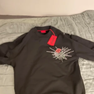 Svart sweatshirt med ros från Hugo - Cool svart sweatshirt från Hugo med stor tryckt ros och taggtrådsmotiv på både fram- och baksida. Tröjan har rund hals och lång ärm, perfekt för dig som vill sticka ut med en unik design. Snygg streetstyle-vibe och riktigt skön passform.